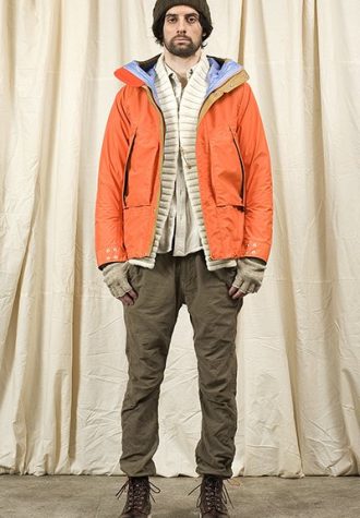 pánská hnědá čepice, oranžová bunda, bílý svetr a hnědé kalhoty Nonnative